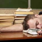 Struggling student resting head on open book with scattered books and glasses on desk, classroom setting, after long study session, fatigue and exhaustion, academic workload burnout, education, learning fatigue.