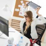 Frustrated woman in an office holding a broken computer monitor above her head, sitting at a desk with a computer, water bottle, and office supplies, expressing workplace stress and technical issues.