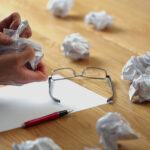 Crumpled paper balls on wooden desk with glasses, pen, and blank sheet, representing brainstorming, frustration, or planning concepts.